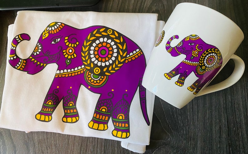May include: A white t-shirt and a white mug with a purple elephant design. The elephant is decorated with intricate patterns in orange, yellow, and green.
