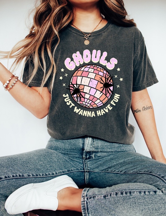May include: Dark gray t-shirt with the words "Ghouls just wanna have fun" in a pink and orange design. The graphic includes a disco ball with spiders and stars. The shirt is a casual, unisex style.