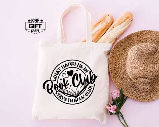 May include: White canvas tote bag with black handles and a black circle design that says "What Happens in Book Club Stays in Book Club" with a stylized open book and two hearts.