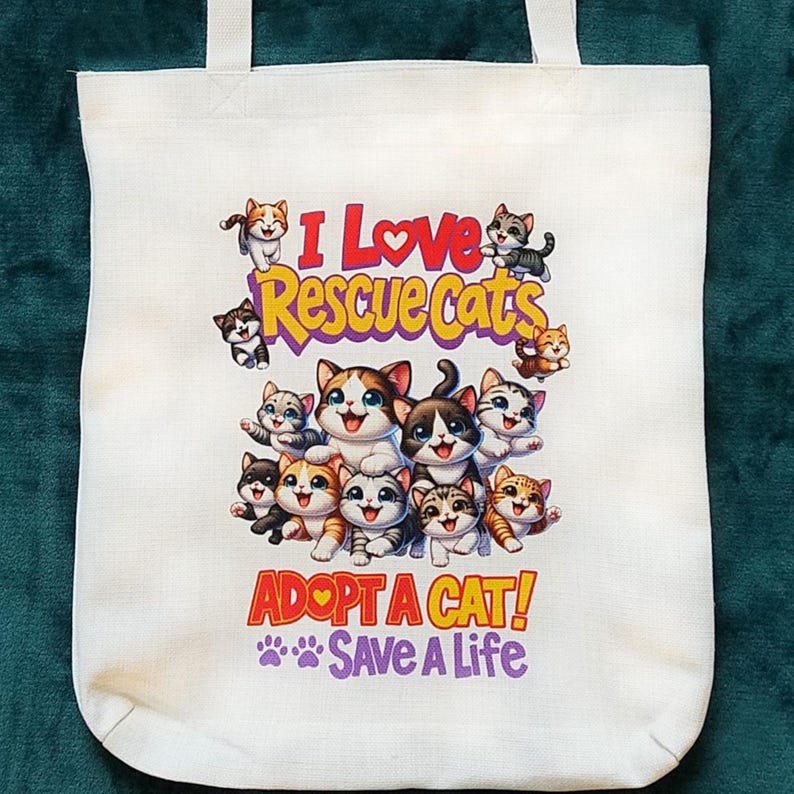May include: White canvas tote bag featuring a colorful graphic of several cartoon cats. The text "I Love Rescue Cats" is above the cats, and "Adopt a Cat! Save a Life" is below. The bag has two white handles.