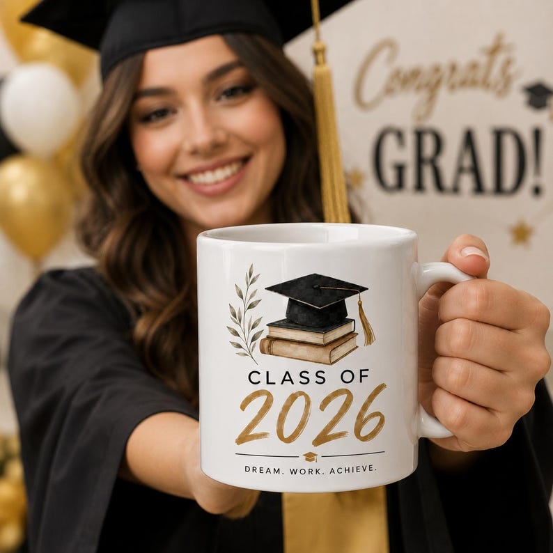 May include: White ceramic mug featuring a graduation cap, books, and botanical elements. The mug reads "Class of 2026" in gold lettering, with the phrase "Dream. Work. Achieve." below. A graduation-themed gift.