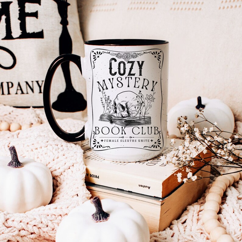 May include: White ceramic mug with a black rim and handle. The mug has a black and white graphic design with the text "Cozy Mystery Book Club - Female Sleuths Unite".